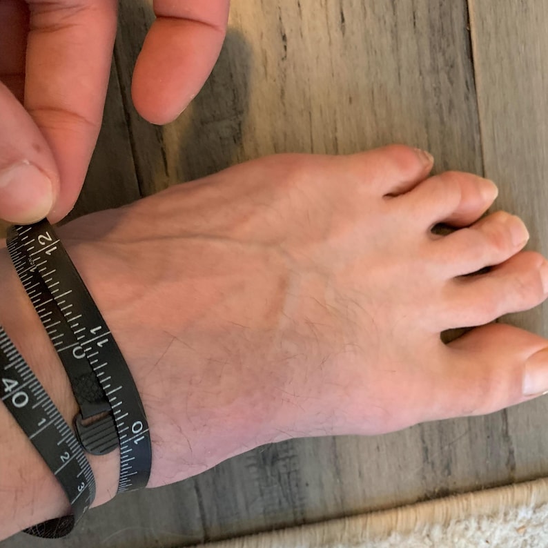 May include: A black measuring tape wrapped around an ankle, showing a measurement of approximately 10 inches. The tape is marked with both inch and centimeter measurements. The foot is resting on a light-colored wooden surface.