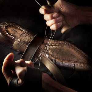May include: A close-up shot of a shoemaker's hands meticulously stitching a leather shoe. The shoe is brown with visible stitching and a leather strap. The shoemaker is using a needle and thread to sew the shoe together.