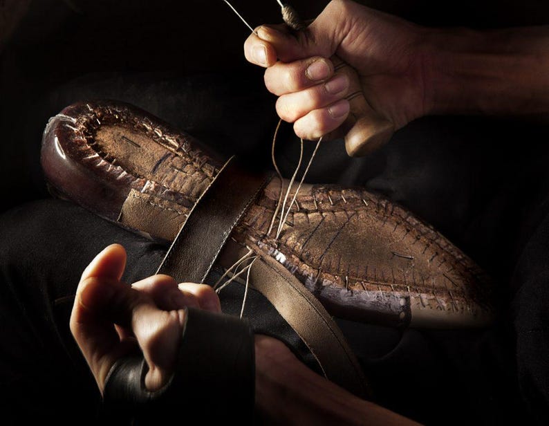 May include: A close-up shot of a shoemaker's hands meticulously stitching a leather shoe. The shoe is brown with visible stitching and a leather strap. The shoemaker is using a needle and thread to sew the shoe together.