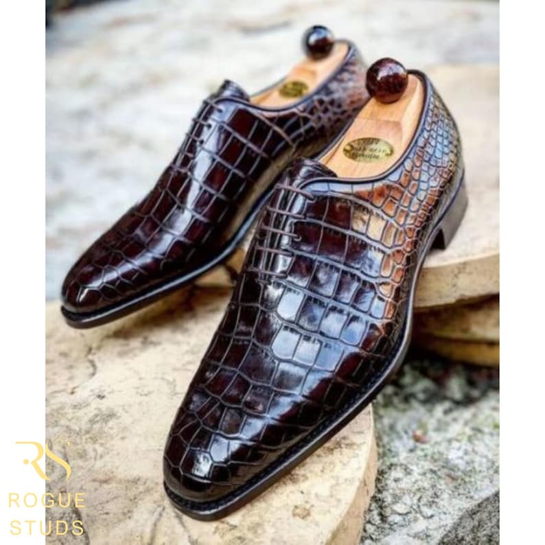 A pair of dark brown, patent leather dress shoes with a crocodile skin pattern. The shoes have a classic design with a pointed toe and a low heel. High quality handmade leather shoes, durable soles and refined craftsmanship