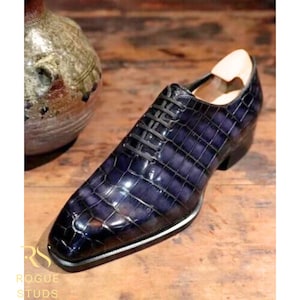 May include: A single, dark blue leather dress shoe with a textured, crocodile-like pattern. The shoe has black laces, a white and silver trim, and a wooden shoe form inside. The shoe is on a wooden surface.