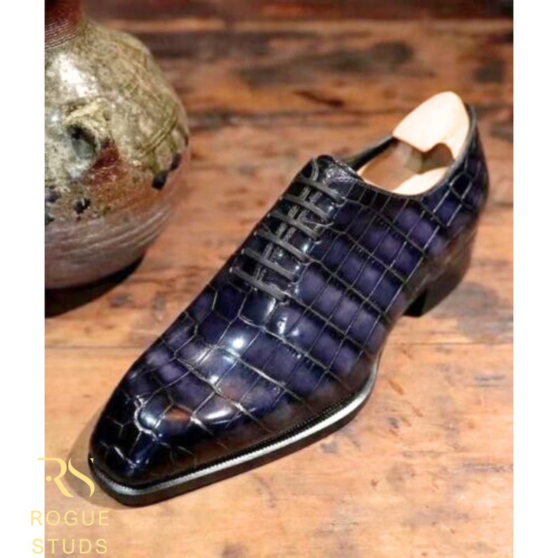May include: A single, dark blue leather dress shoe with a textured, crocodile-like pattern. The shoe has black laces, a white and silver trim, and a wooden shoe form inside. The shoe is on a wooden surface.