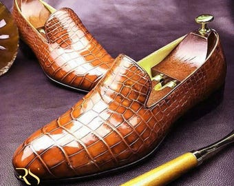 May include: A pair of brown leather loafers with a crocodile skin texture. The shoes have a classic slip-on design and are displayed with shoe trees inside. The image also includes a smoking pipe and the text "ROGUE STUDS".