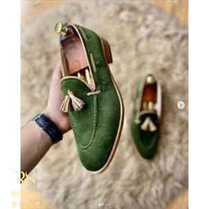 May include: Green velvet loafers with tassel detail. The shoes have a classic design with a rounded toe and a low heel. The loafers are displayed on a textured surface.