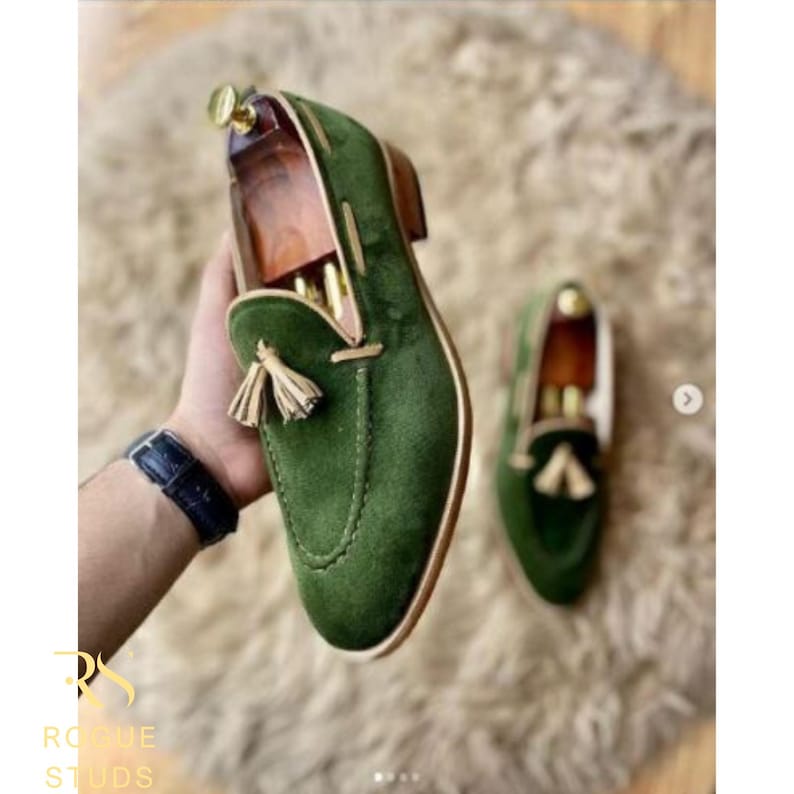 May include: Green velvet loafers with tassel detail. The shoes have a classic design with a rounded toe and a low heel. The loafers are displayed on a textured surface.