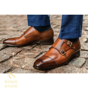 May include: A pair of brown leather double monk strap shoes with a polished finish. The shoes feature a cap toe design and gold-colored buckles. The image shows the shoes on a cobblestone surface, with blue pants visible.