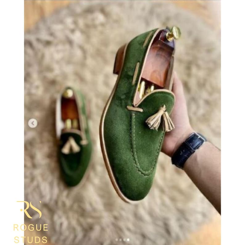 May include: Green suede loafers with a tassel detail. The shoes have a rounded toe and a low heel. The image also shows a second shoe in the background. The shoes are on a fluffy, light-colored surface.