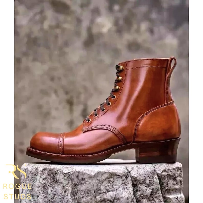 May include: A brown leather boot with a cap toe and lace-up closure. The boot has a stacked heel and a pull tab at the back. The boot is sitting on a stone surface. The text "ROGUE STUDS" is visible in the lower left corner.