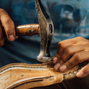 May include: A close-up shot of a shoemaker using a hammer to work on a leather shoe. The hammer is silver with a wooden handle. The shoe is brown and beige, with visible stitching and metal studs.