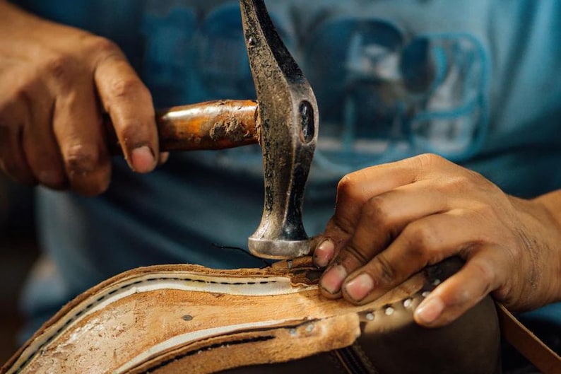May include: A close-up shot of a shoemaker using a hammer to work on a leather shoe. The hammer is silver with a wooden handle. The shoe is brown and beige, with visible stitching and metal studs.