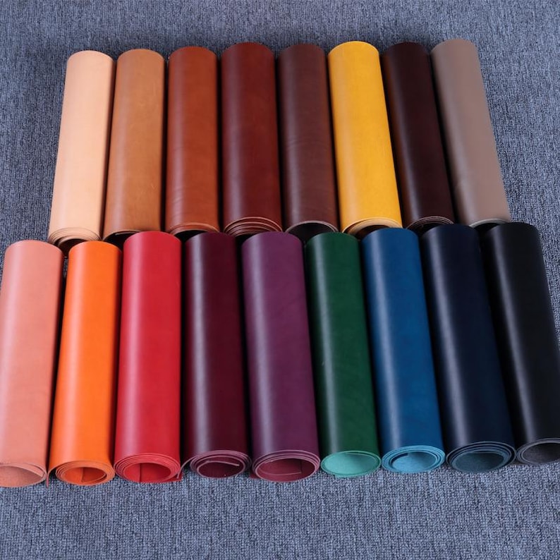 May include: A collection of rolled leather sheets in various colors, including tan, brown, red, purple, green, blue, and black. The leather appears smooth and is arranged on a gray surface. The rolls are tightly wound, showcasing the different hues available.