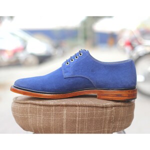 May include: A single blue suede dress shoe with a brown leather sole and heel. The shoe has a classic lace-up design with dark blue laces and is displayed on a brown patterned cushion.