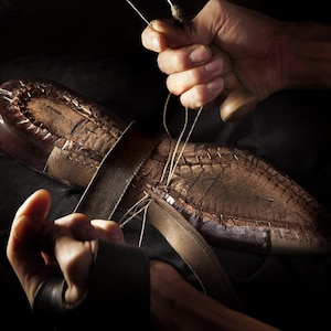 May include: A close-up shot of a shoemaker's hands meticulously stitching a leather shoe. The shoe is brown with visible stitching and a leather strap. The shoemaker uses a needle and thread to sew the shoe together.