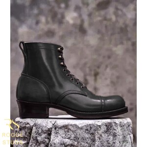 May include: A dark green leather lace-up boot with a cap toe featuring decorative perforations, a stacked heel, and a pull tab at the top of the shaft. The boot has dark eyelets and matching laces, showcasing a smooth finish and structured design.