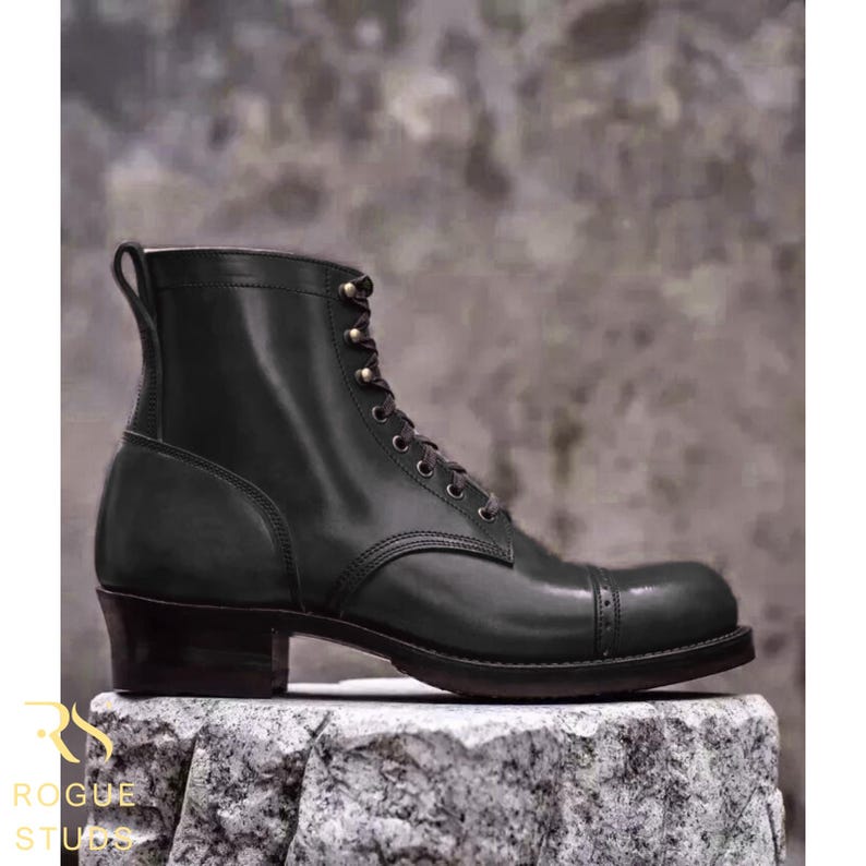 May include: A dark green leather lace-up boot with a cap toe featuring decorative perforations, a stacked heel, and a pull tab at the top of the shaft. The boot has dark eyelets and matching laces, showcasing a smooth finish and structured design.
