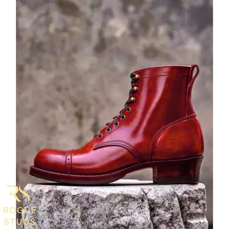 May include: A polished, burgundy leather boot with a cap toe and lace-up closure. The boot features a stacked heel and brass eyelets. The brand name "ROGUE STUDS" is visible in the lower left corner. The boot is approximately 6 inches tall.