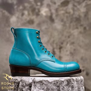 May include: A vibrant turquoise leather boot featuring a cap toe with decorative perforations, brass eyelets, and a dark brown stacked heel. The side of the boot displays a pull tab at the collar and the text ROGUE STUDS in gold lettering.