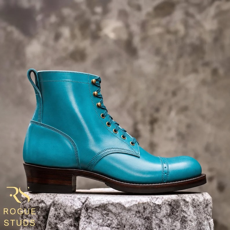 May include: A vibrant turquoise leather boot featuring a cap toe with decorative perforations, brass eyelets, and a dark brown stacked heel. The side of the boot displays a pull tab at the collar and the text ROGUE STUDS in gold lettering.