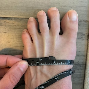 May include: A foot is being measured with a black measuring tape. The tape is wrapped around the foot, showing the width measurement. The foot is on a light brown wooden surface.