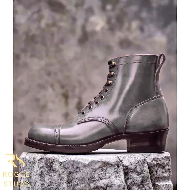 May include: A gray leather boot with a cap toe and lace-up closure. The boot has a stacked heel and is displayed on a stone surface. The brand name "ROGUE STUDS" is visible in the lower left corner.