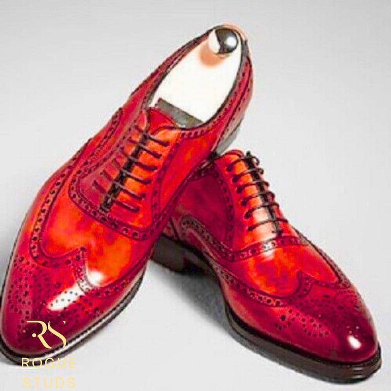 May include: A pair of red leather dress shoes with brogue detailing and black laces. The shoes have a polished finish and a classic wingtip design. The image shows the shoes from a slightly elevated angle, highlighting their shape and texture.