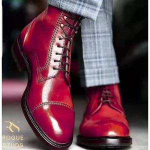 May include: A pair of burgundy leather boots with black laces and a cap toe. The boots have a high-shine finish and are laced up. The boots are part of the Rogue Studs collection.
