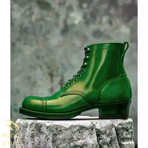May include: A single, glossy green leather boot with a cap toe and lace-up closure. The boot has a stacked heel and a pull tab at the top. The boot is displayed on a textured stone surface.