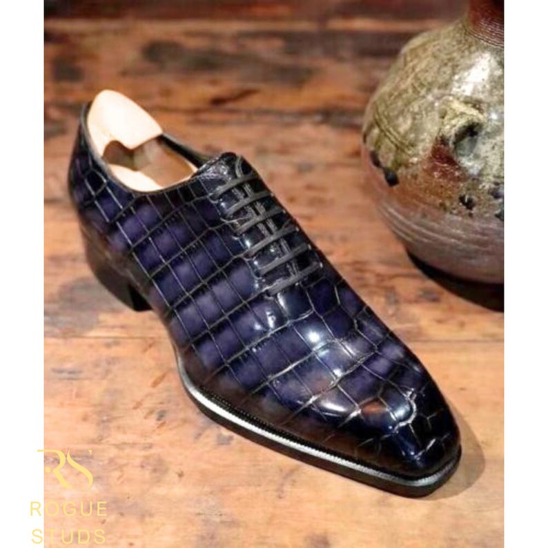 May include: A single, dark blue leather dress shoe with a textured, crocodile-like pattern. The shoe has black laces, a black heel, and a white and silver trim along the sole. The shoe is on a wooden surface.