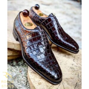 May include: A pair of brown leather dress shoes with a crocodile skin pattern. The shoes have a glossy finish and are displayed with wooden shoe trees inside. The shoes are on a stone surface.
