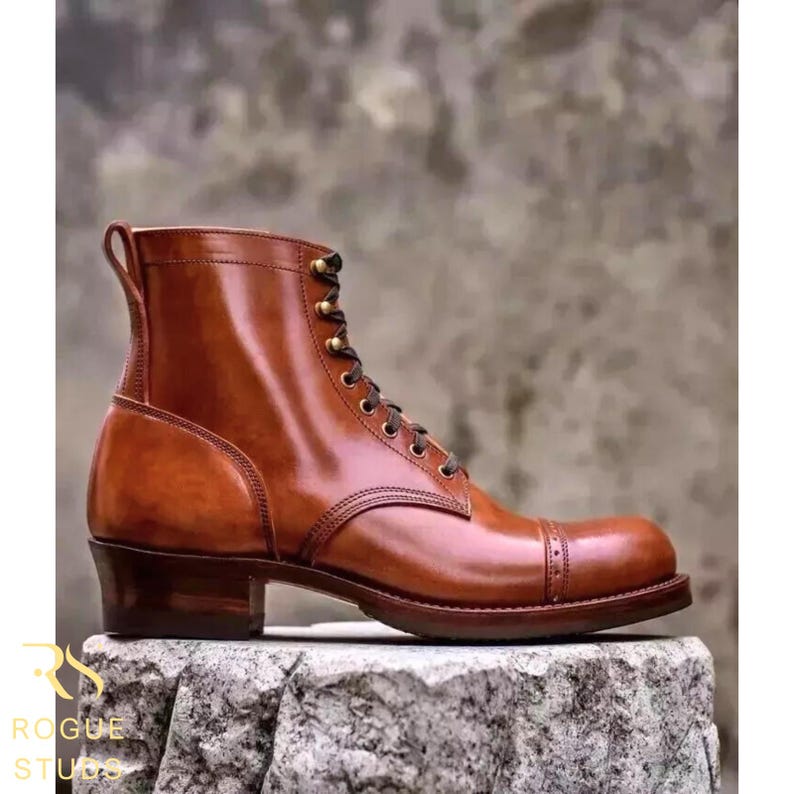 May include: A brown leather boot with a cap toe and lace-up fastening. The boot features a stacked heel and a pull tab. The boot is displayed on a stone surface. The text "ROGUE STUDIO" is visible.