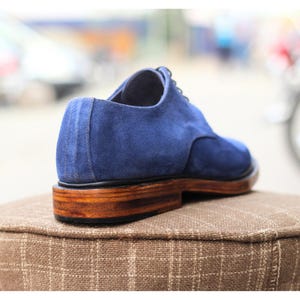 May include: A blue suede oxford shoe with a wooden sole. The shoe has a classic design with a lace-up closure. The heel is approximately 1 inch high. The shoe is sitting on a brown, textured surface.