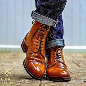 May include: Smart, brown leather boots with black laces, worn with rolled-up denim jeans. The boots have a cap-toe design and a polished finish. The boots are on a stone surface. The text "ROGUE STUDS" is visible in the lower left corner.