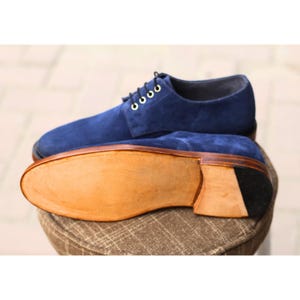 May include: A pair of blue suede dress shoes with a leather sole. The shoes have a classic lace-up design with gold-colored eyelets. The sole is a light brown color with a black heel.