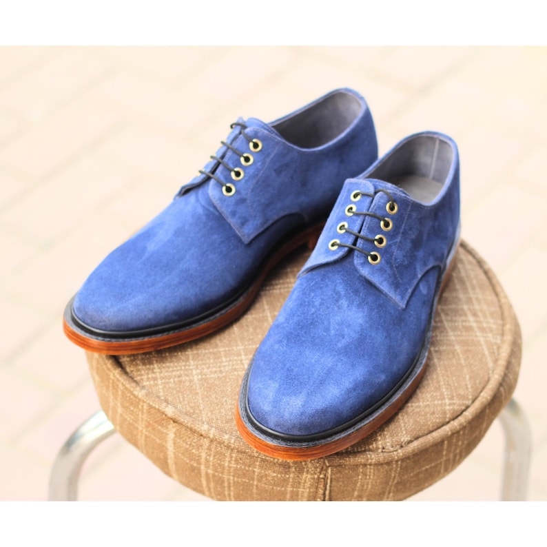 May include: A pair of blue suede dress shoes with black laces and gold eyelets. The shoes have a brown sole and are displayed on a small, round stool with a patterned fabric.