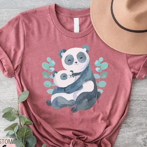 May include: A dusty rose t-shirt featuring a watercolor illustration of two pandas hugging, surrounded by green leaves. The shirt has rolled-up sleeves and a knotted hem. The text "Trendy Shirts" is printed on the bottom left.