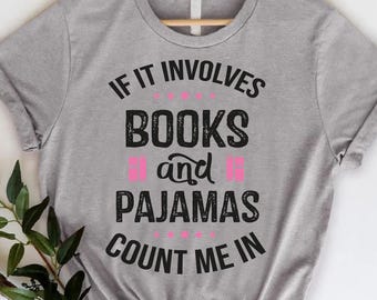 May include: A heather gray t-shirt with the text "IF IT INVOLVES BOOKS and PAJAMAS COUNT ME IN" in black and pink. The shirt is hanging on a wooden hanger against a white background. A leafy branch is in the lower left corner.
