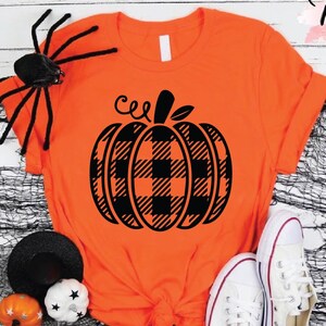May include: Orange t-shirt with a black plaid pumpkin graphic, white sneakers, and Halloween decorations. The shirt has the text "Custom Trendy Shirts" in a script font. A black spider, pumpkins, and a black hat are also present.