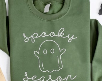 May include: Olive green sweatshirt with the words "spooky season" and a ghost graphic in white. The image includes a tombstone, skull, and black paper butterflies, creating a Halloween theme.