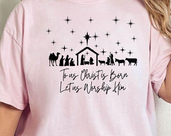 May include: A light pink t-shirt featuring a black graphic of a nativity scene with stars and the text "To us Christ is Born, Let us Worship Him." The shirt is a relaxed fit.