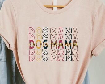 May include: A pink t-shirt with the text "DOG MAMA" repeated in a retro style with colorful accents. The text is layered with a yellow, red, and blue pattern.