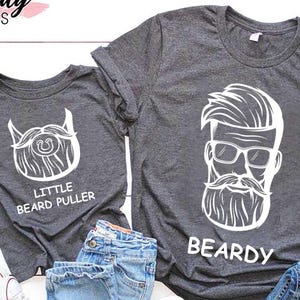 May include: Two dark gray t-shirts with white graphic beard designs and text. One shirt says "Little Beard Puller" and the other says "Beardy". Blue jeans and white sneakers are also in the image.