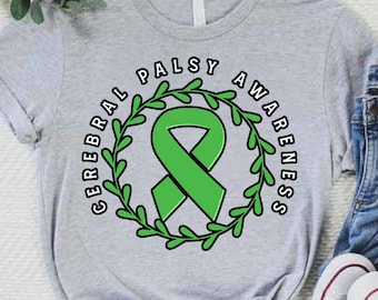May include: A light gray t-shirt with the words "CEREBRAL PALSY AWARENESS" encircling a green ribbon and a green leafy wreath. The shirt is paired with blue jeans and white sneakers.