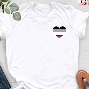 May include: White t-shirt with a heart-shaped design in black, gray, and purple, representing the asexual pride flag. The shirt is part of a clothing collection, with the text "Custom Trendy Shirts" in the upper right corner. The shirt is paired with blue jeans and brown sandals.