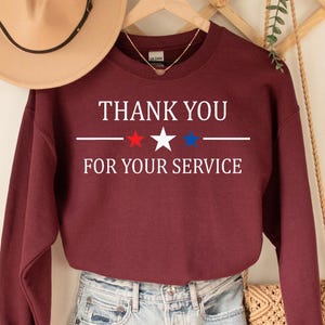 May include: Burgundy sweatshirt with the text "THANK YOU FOR YOUR SERVICE" in white, with red and blue star accents. The sweatshirt is paired with denim shorts and a tan hat. A wooden hanger and decorative wall piece are also visible.