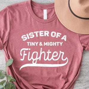 May include: A maroon t-shirt with the text "Sister of a Tiny & Mighty Fighter" in white. The shirt is made of a soft, comfortable material and is perfect for everyday wear.