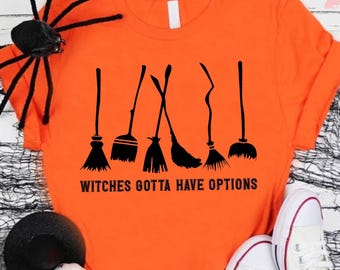 May include: Orange t-shirt with the text "Witches Gotta Have Options" and a graphic of several black witch brooms. The shirt is styled with white sneakers, a black spider, and Halloween decorations.