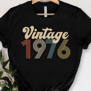 May include: Black t-shirt with the text "Vintage 1976" in a retro font. The numbers are in red, blue, and yellow. The shirt is hanging on a wooden hanger.