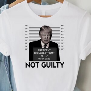 May include: White t-shirt featuring a mugshot-style image of Donald J. Trump with the text "NOT GUILTY." The image includes a height chart and a sign with the text "PRESIDENT DONALD J TRUMP 45-47 04 04 2023."