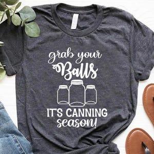 May include: Dark gray t-shirt with the text "grab your Balls, it's canning season!" in white. The shirt is paired with blue jeans and brown sandals with black straps. Green leafy sprigs are in the upper left corner.