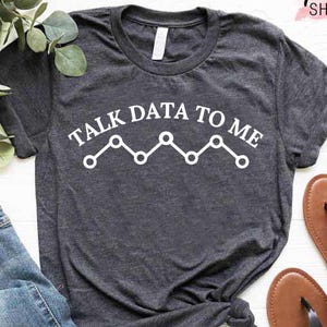 May include: Dark gray t-shirt with the text "TALK DATA TO ME" in white, along with a white line graph design. The shirt is paired with blue jeans and brown sandals with black straps. The words "CUSTOM Trendy SHIRTS" are in the top right corner.